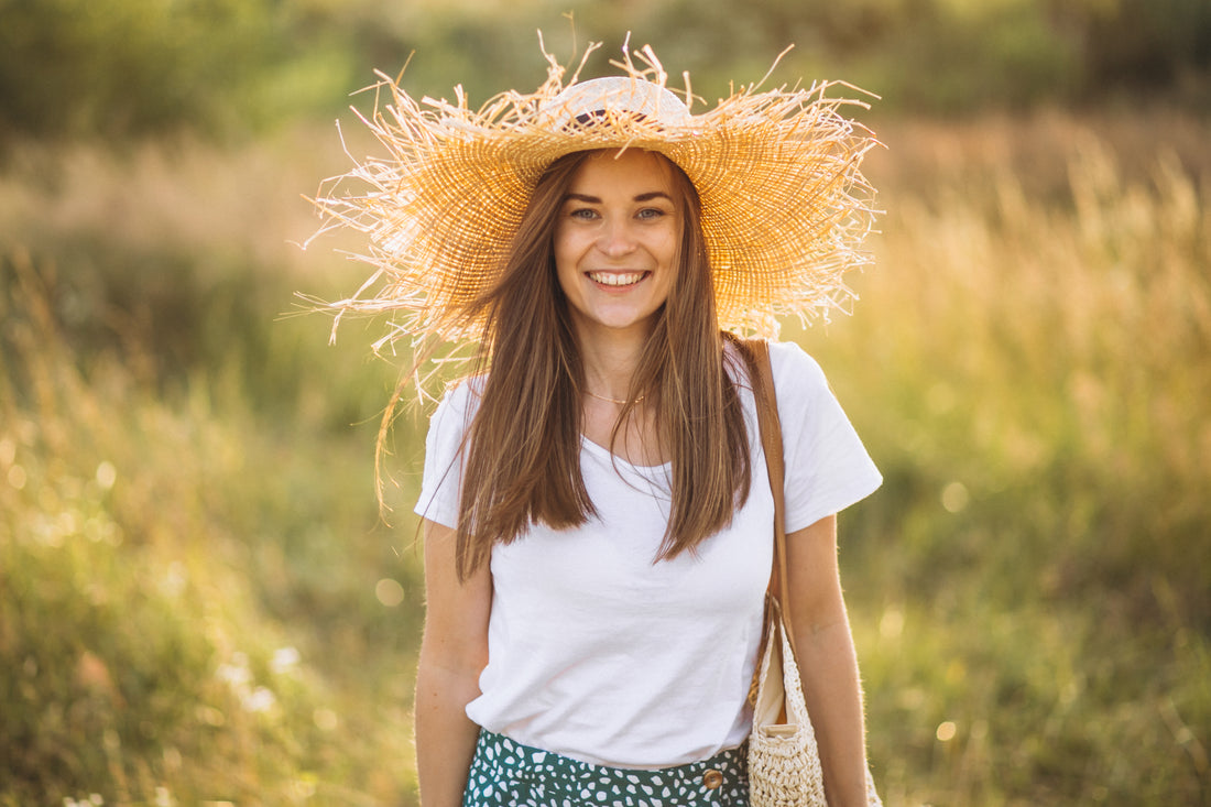 A smiling young woman stands in a vibrant field of flowers, wearing a white t-shirt and a floral skirt. Her radiant straw hat complements her joyful expression, showcasing the harmony between nature-inspired style and positivity.