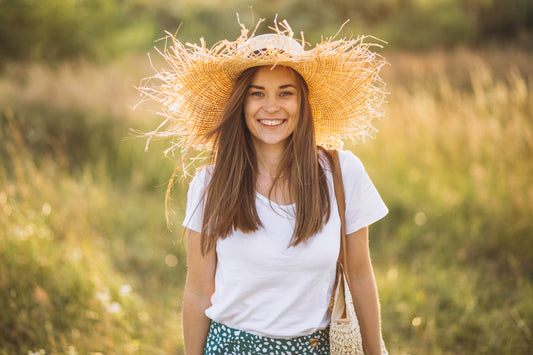 A smiling young woman stands in a vibrant field of flowers, wearing a white t-shirt and a floral skirt. Her radiant straw hat complements her joyful expression, showcasing the harmony between nature-inspired style and positivity.